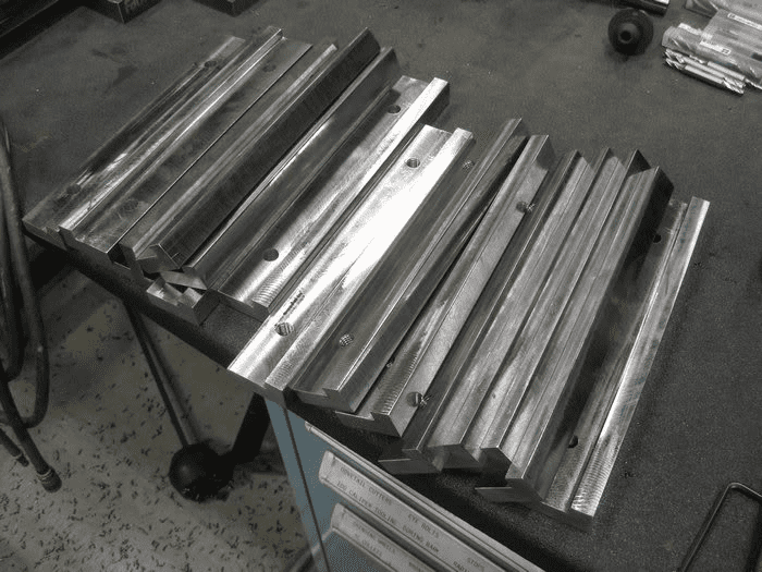 Metal parts arranged on a table, featuring rectangular shapes with rough texture, small holes or protrusions. Durable material likely steel or aluminum.