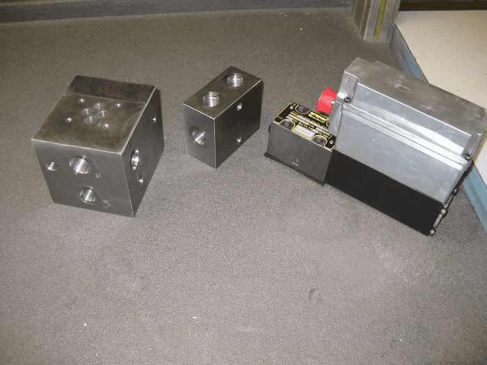 Metal parts on gray surface, with square blocks, circular holes, and a small rectangular block with a red lever.