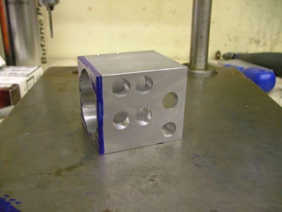 Silver-colored aluminum cube with circular holes on each side, sitting on a dark gray metal workbench.