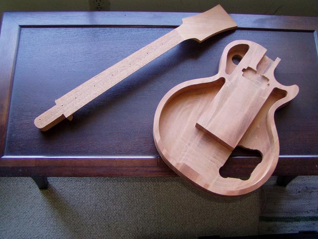 A partially constructed wooden guitar body and neck on a dark wood table, made from light-colored hardwood with visible grain patterns.