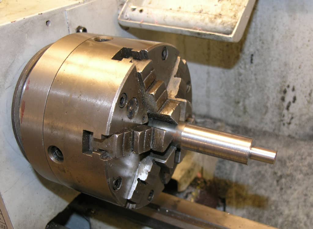 A cylindrical metal component with a central rod protruding from its side, featuring small holes along its circumference, attached to a wall.