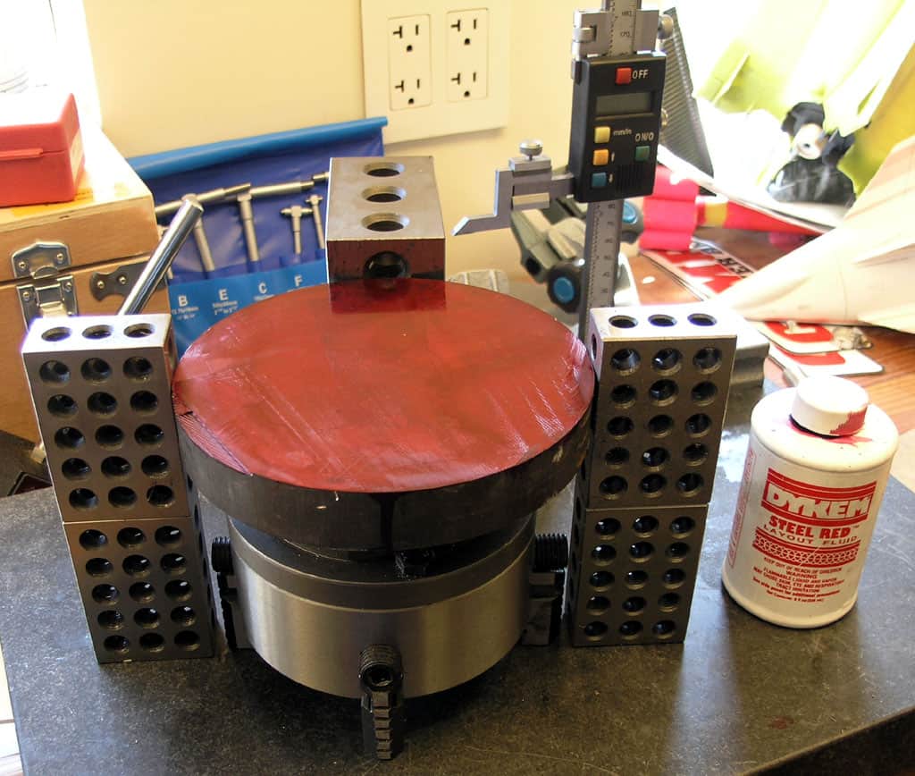 Red circular metal surface on base with holes, surrounded by metal blocks with holes, likely a lathe or milling machine component.