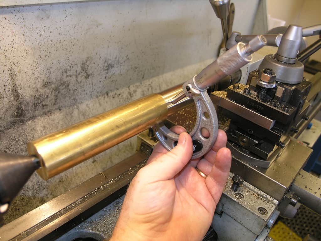 A person's hand holds a small metal object in front of a lathe machine with scattered tools and parts on the workbench.