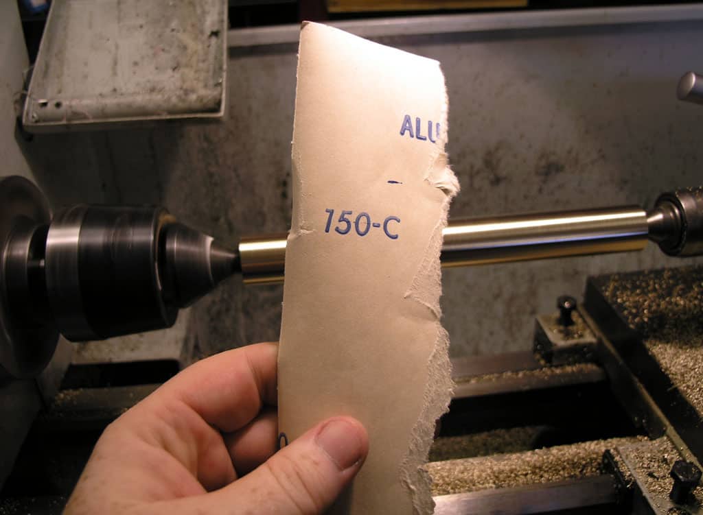 A hand holds sandpaper labeled "150-C ALU" in a machine shop or metal fabrication setting with background machinery components.
