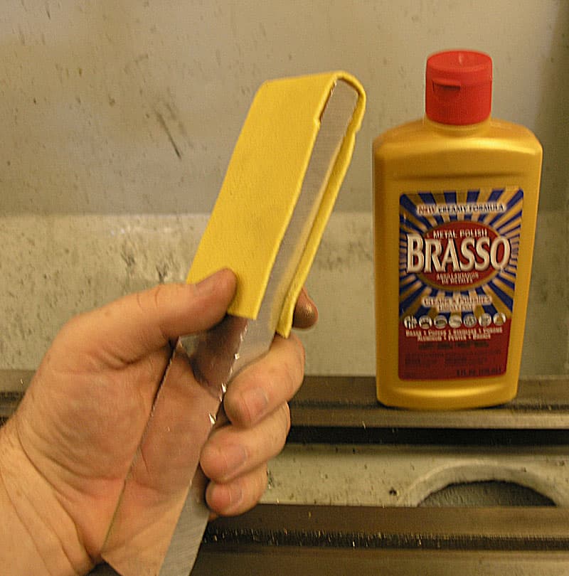 A hand holding a yellow sponge near Brasso polish and a metal tool on a workbench with a gray wall in the background.