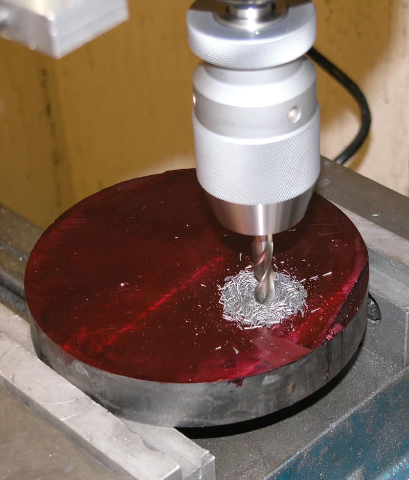 A drill bit creates a hole in a red, possibly metallic or plastic, object on a workbench with a drill press.