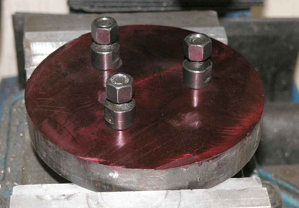 Round metal object with red coating and four bolts on top, situated in an industrial setting.