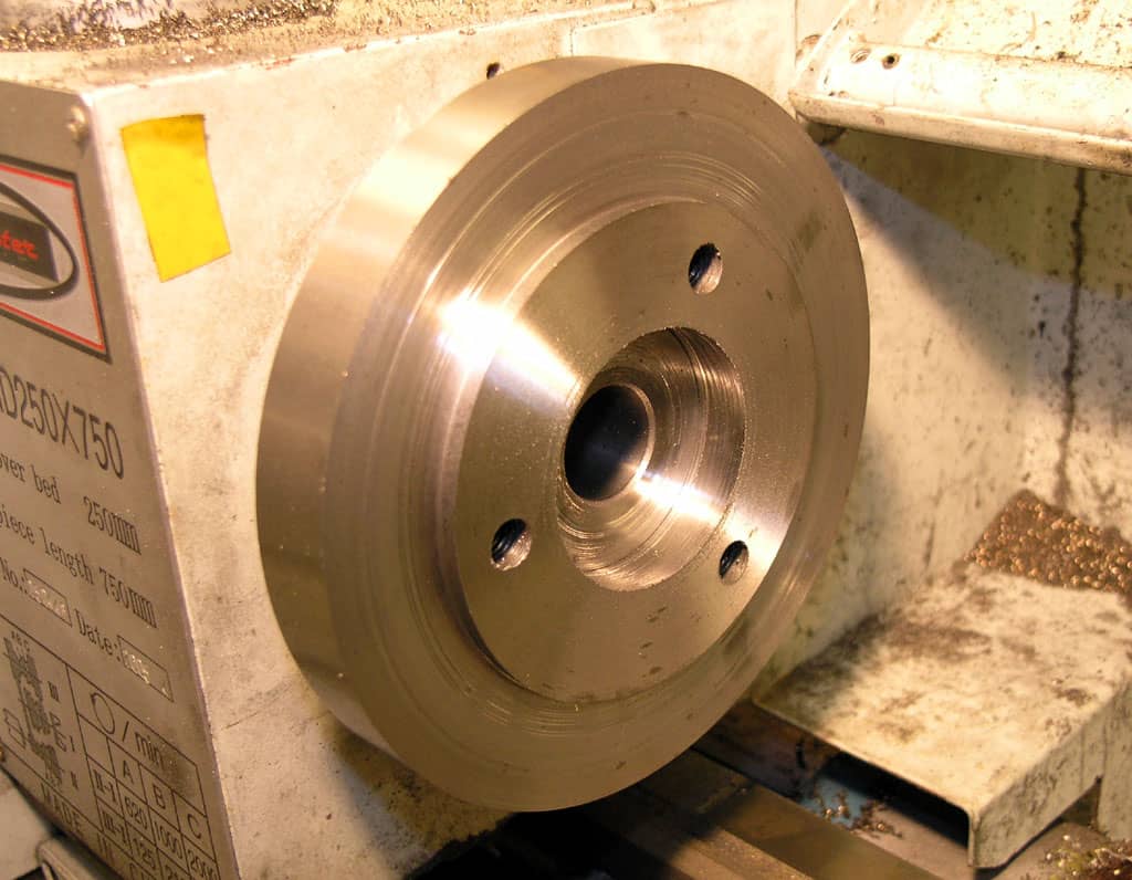 A large metal wheel with four holes, shiny metal finish and visible ridges.
