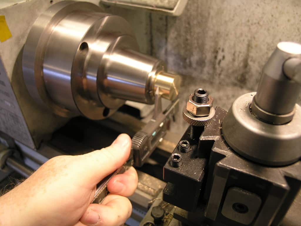 A person's hand adjusts a small black knob on a metal lathe in a busy machine shop.