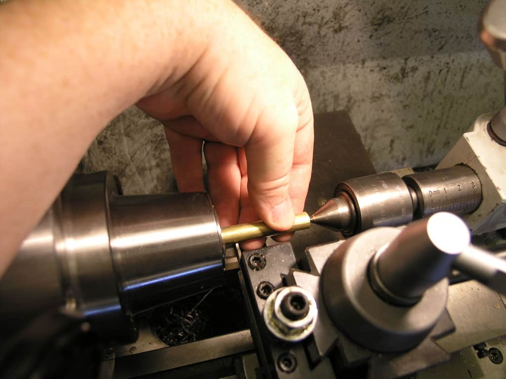 A person operates a metal lathe with one hand holding a brass rod.