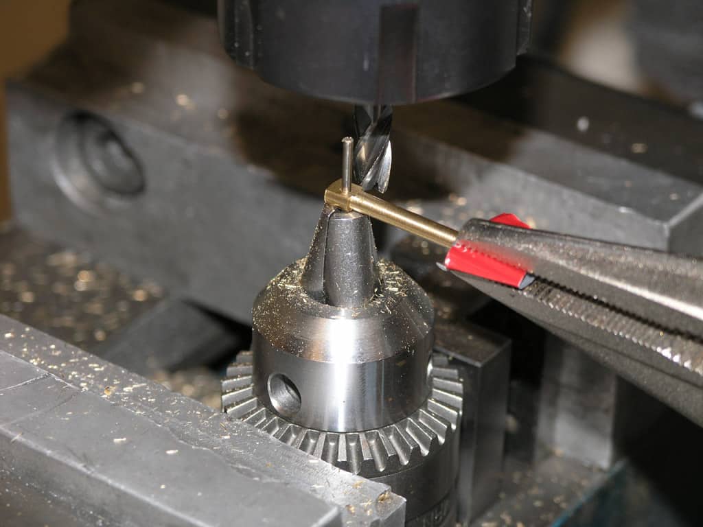 A drill bit in a drill press, held by pliers in a metal fabrication workshop.