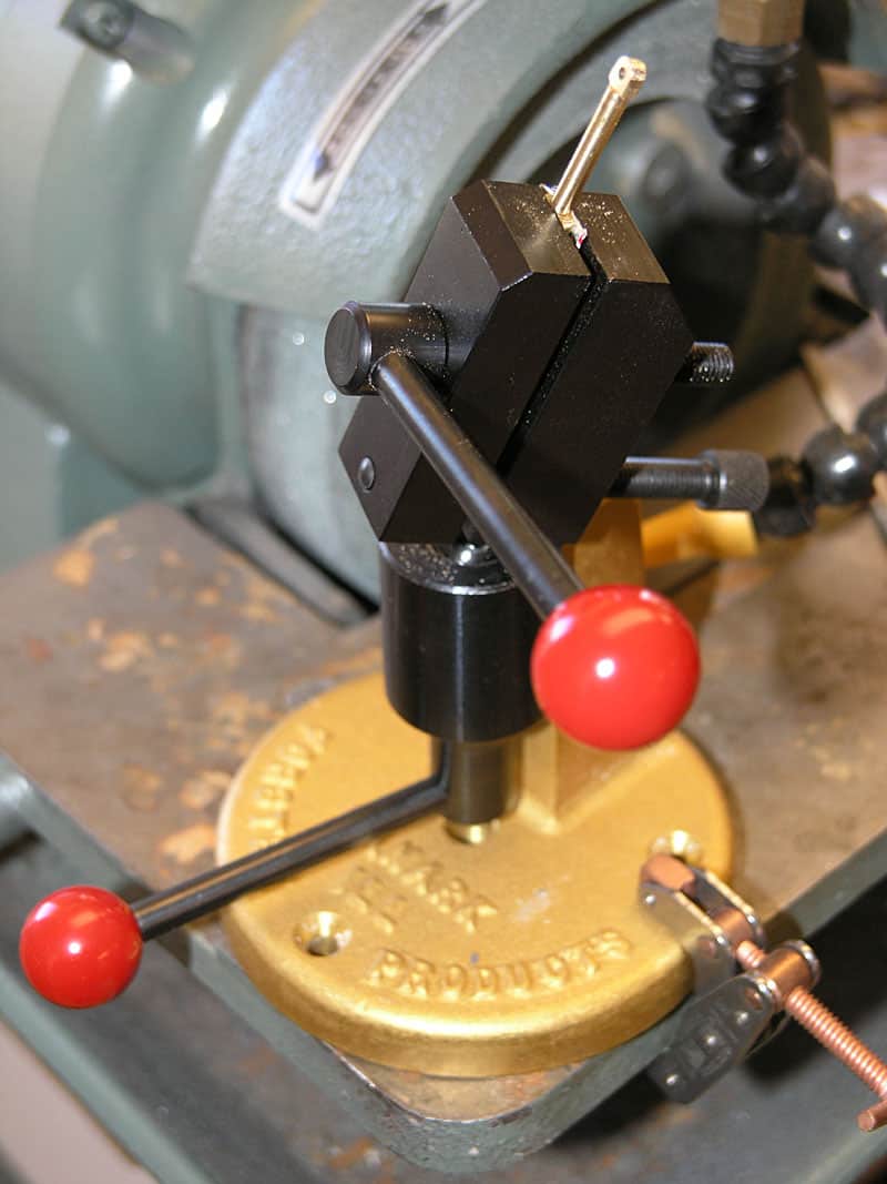 Small black metal vise with red knobs holding a brass rod on a round gold base in a workshop setting.