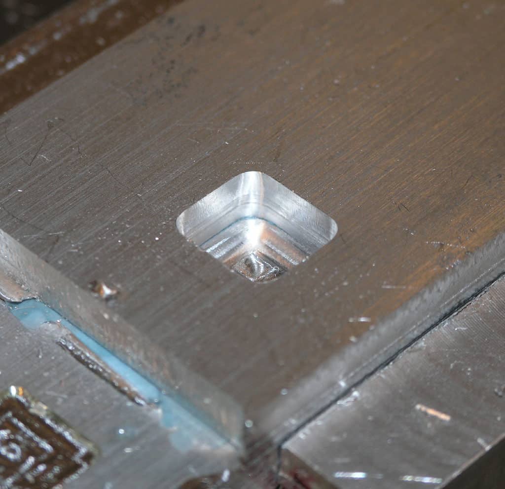 Square metal object with recessed area, likely from machining or metal fabrication process.