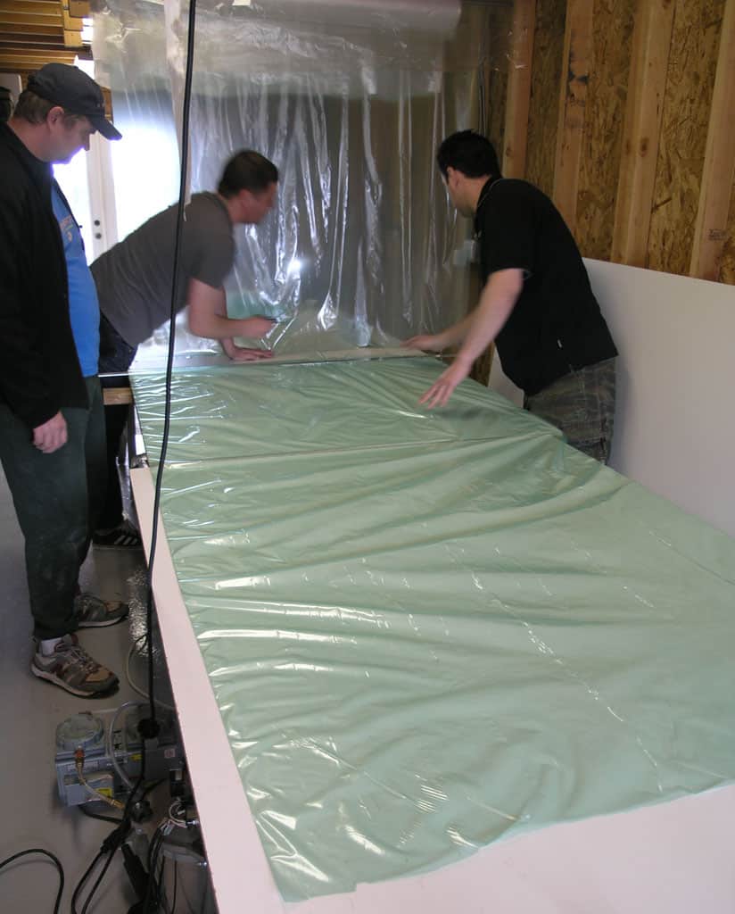 Men laying down a large light green sheet of thin, flexible plastic material on a white table in a workshop.