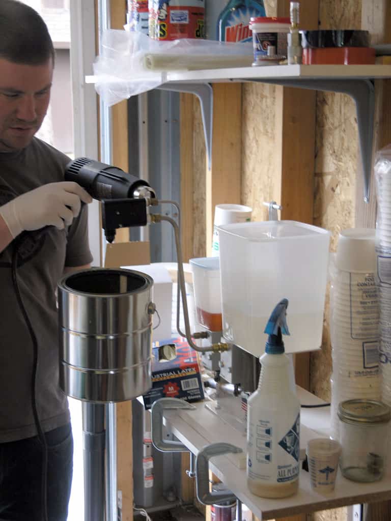 Man in gray shirt and white gloves standing next to metal container with drill.