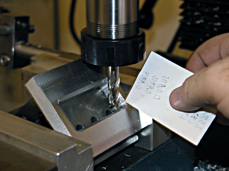 Person holding white card above metal drill press or milling machine in a workshop setting.