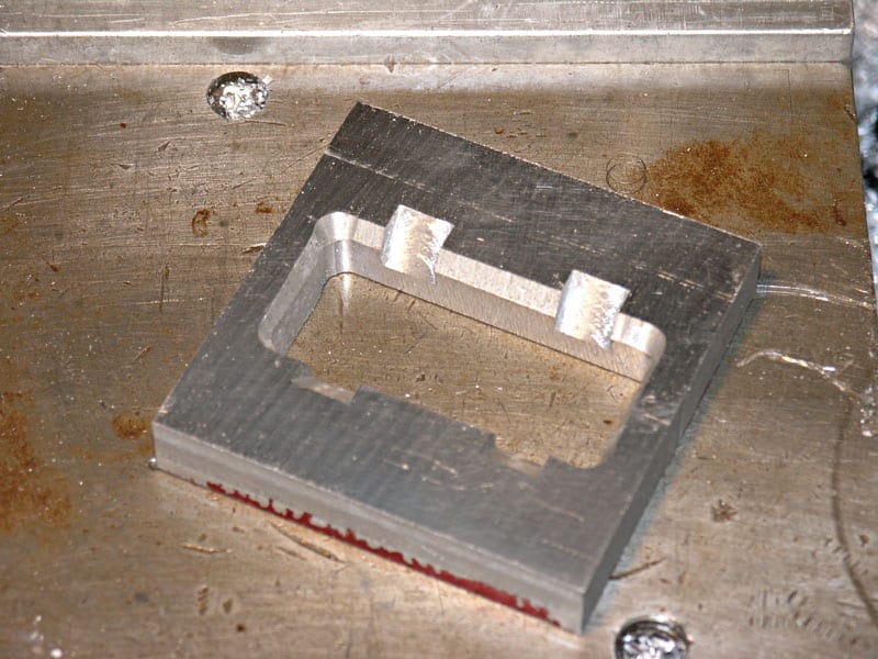 A square metal object with a central rectangular recessed area, featuring a rough, unfinished aluminium-like surface.
