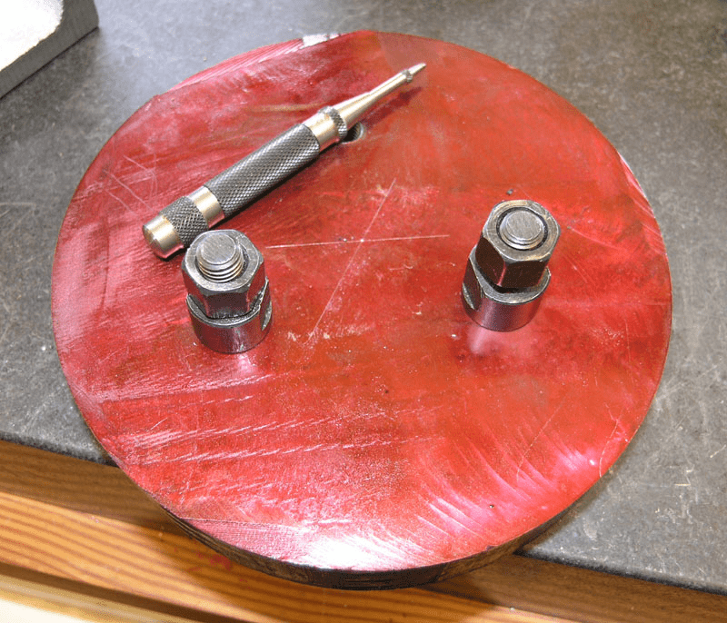 Round red metal tool with three silver bolts, resting on gray surface.