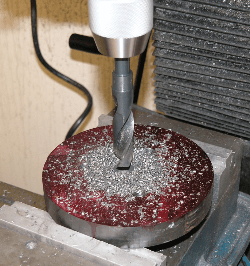 A hand drill or drill press in use, with a large metal piece being drilled through.