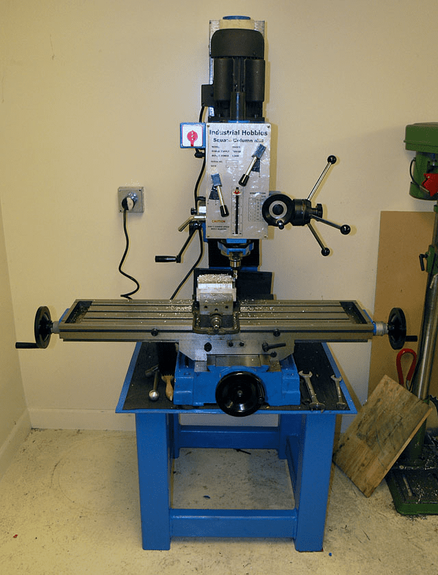 Blue industrial milling machine with controls and electrical outlet in front of white wall.