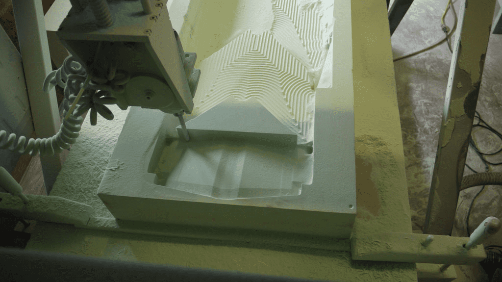 Large-scale 3D milling machine in operation, shaping a rectangular piece of material with ridges and grooves.