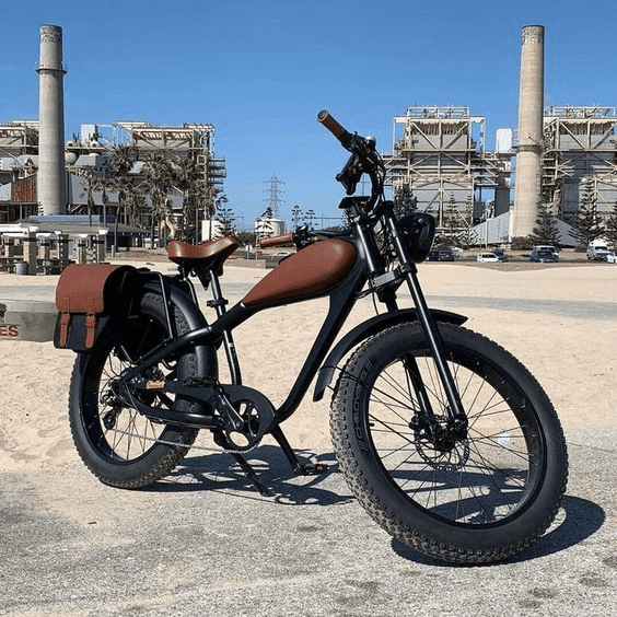 A fat-tire electric bicycle with brown leather seat and saddlebags parked on a beach in front of an industrial complex.