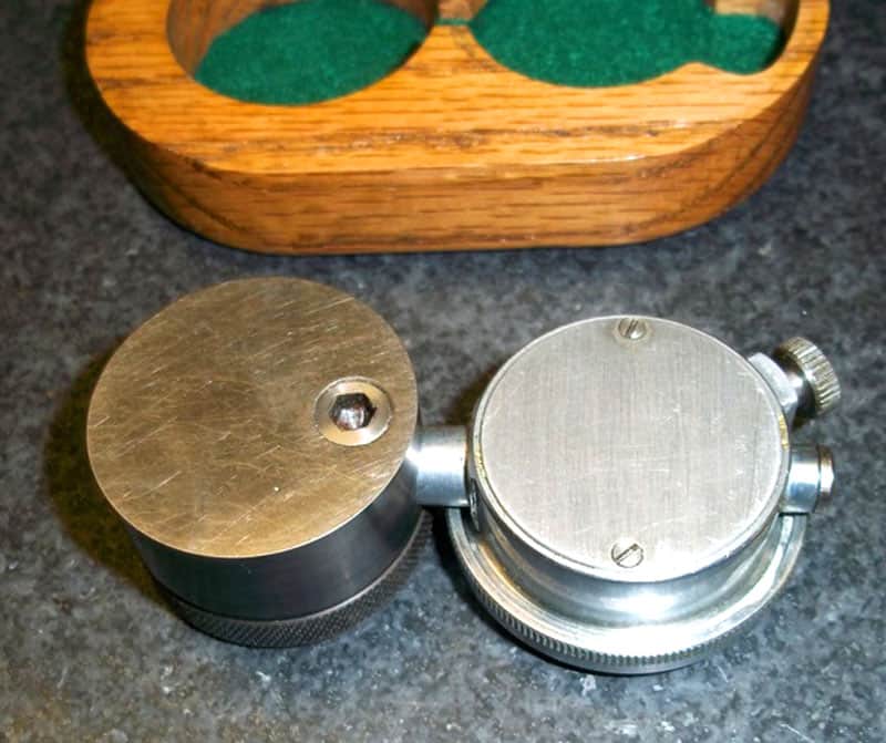 Two metal objects with rounded edges and screw-like protrusions on a gray surface next to a wooden box.