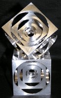 Metallic object with square base and diamond-shaped top, featuring circular cutouts on sides, made of metal with shiny surface.