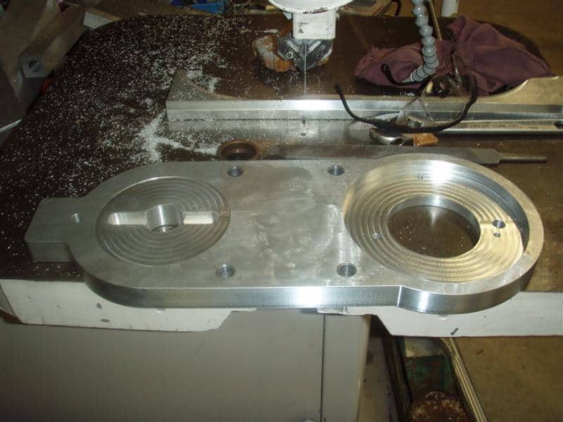 A large aluminum part with two circular recesses sits on a metal workbench in a cluttered machine shop workshop.