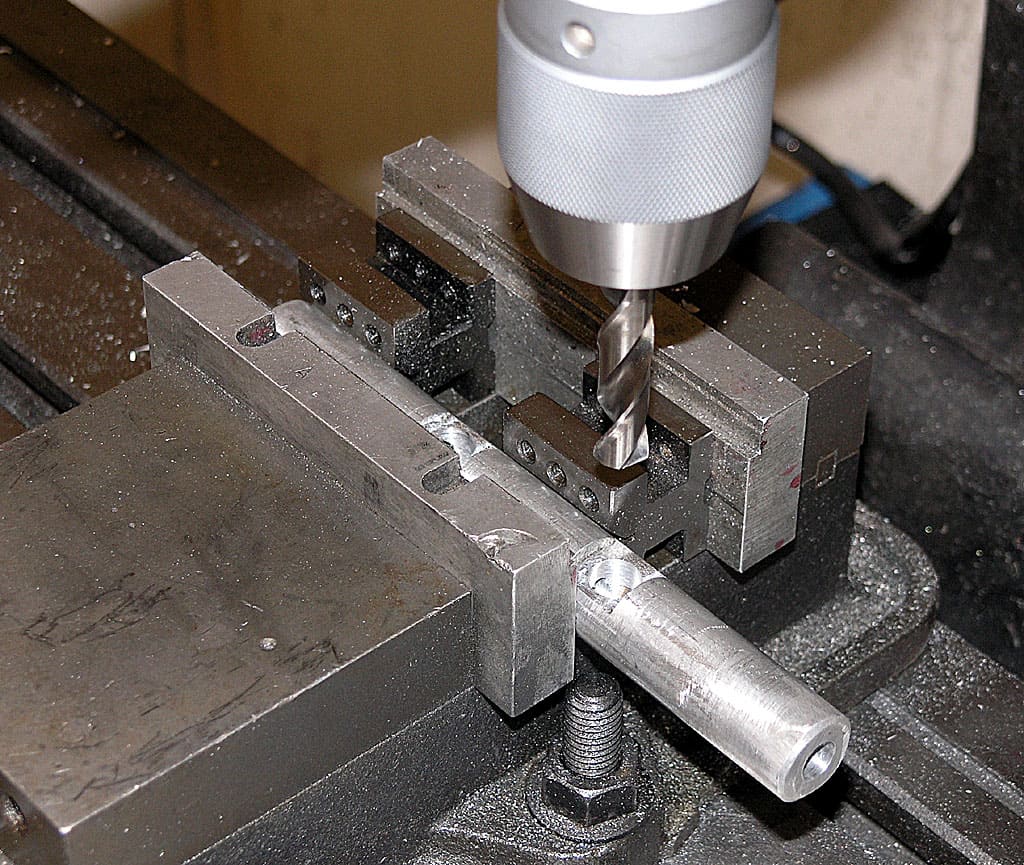 Silver drill bit with spiral flute attached to drill press chuck over metal workpiece on a vise in a machine shop setting.