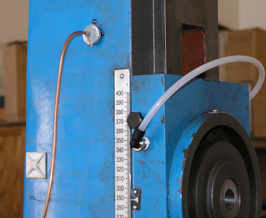 Industrial device with blue body, copper tube, white gauge, and black knob in a workshop or laboratory setting.