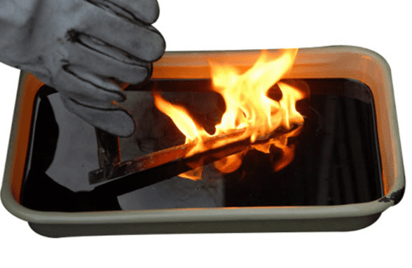 A rectangular metal container holds dark liquid as a gloved hand places a lit match nearby.
