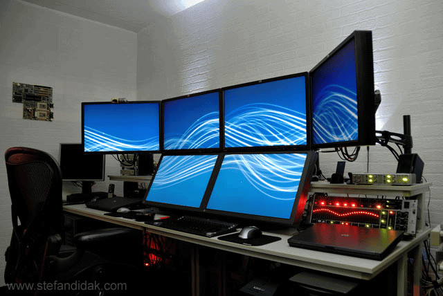 Four computer monitors on a wall with keyboard, mouse, and cables on a desk in front of a white brick wall.
