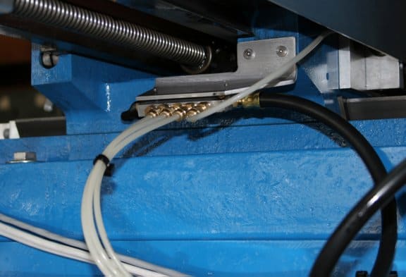 Metal object with blue background, silver component on left, textured surface, black hose, white wires, gray spring, small hole on right.