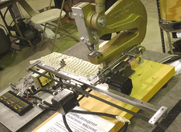 Green metal benchtop punching machine with yellow platform, wires, and components on gray workbench.
