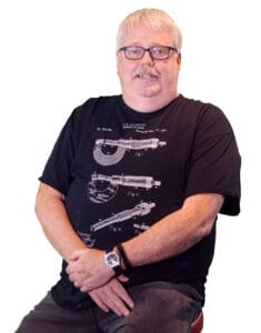 A middle-aged man with short white hair, wearing a black t-shirt and dark pants, sits with arms crossed and a smile.