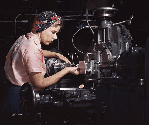 A woman operates a large metal lathe, wearing a black and red floral headscarf.