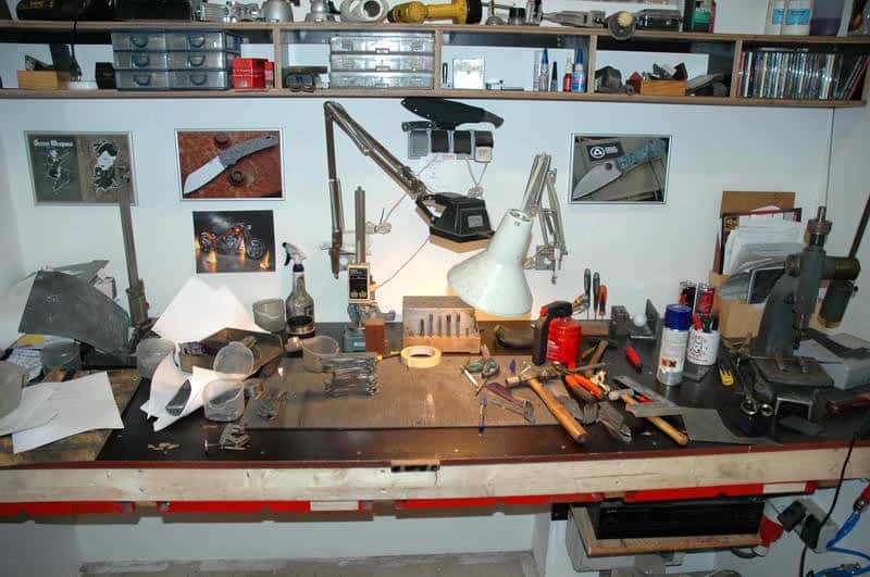 A cluttered workbench in a well-lit workshop or garage with various dark wood tools and equipment, including a vice, drill press, and lathe.