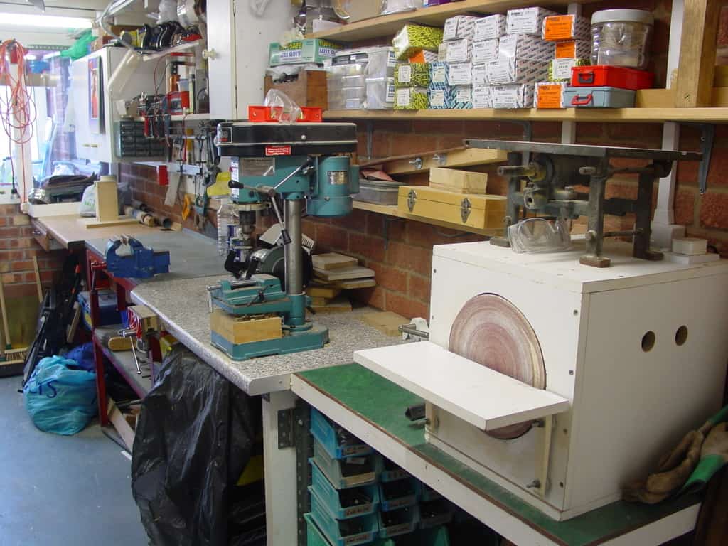 Well-equipped metalworking workshop with drill press, bandsaw, and sanding drum on wooden workbenches.