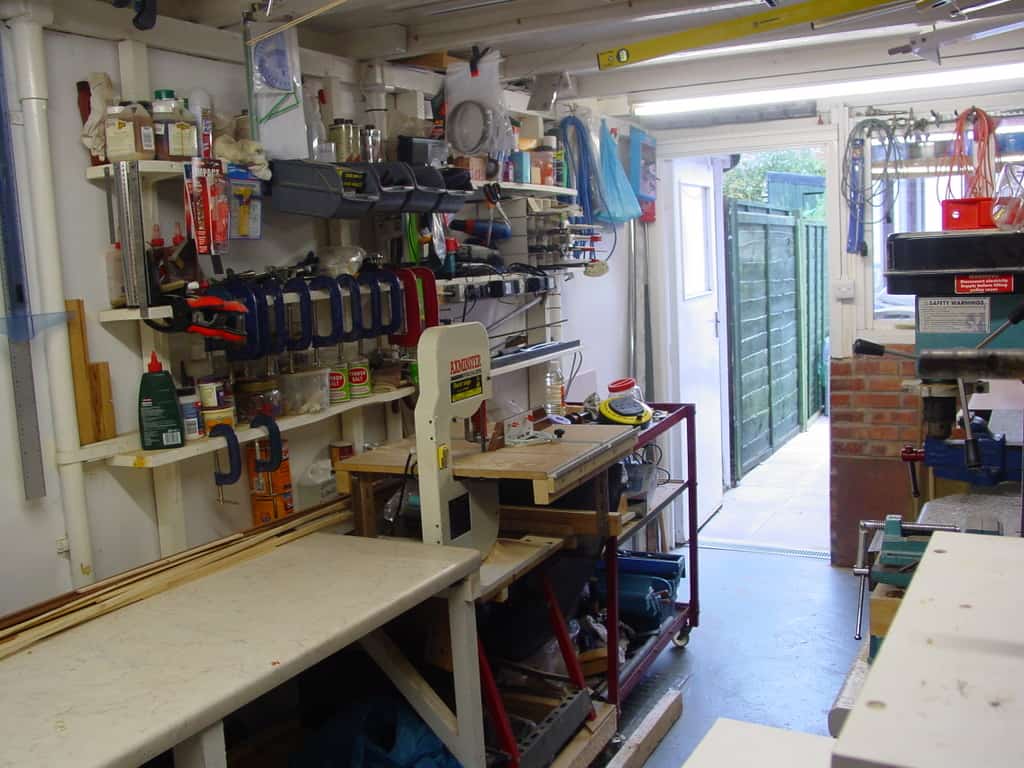 Cluttered workshop with tools, equipment, shelving unit, workbenches, and machinery in a well-lit room.