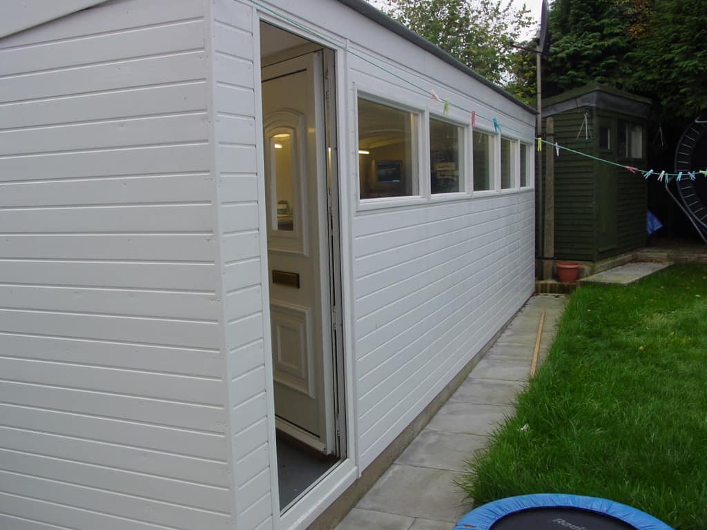 White shed with open door in backyard setting featuring lawn, trampolines, and surrounding outbuildings and trees.