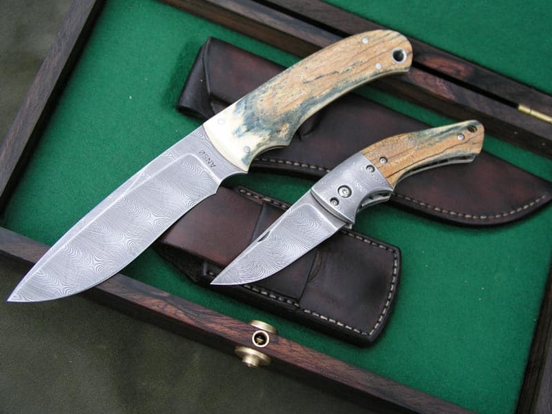 Two knives with intricate patterns on blades and wooden handles in a green felt-lined box.