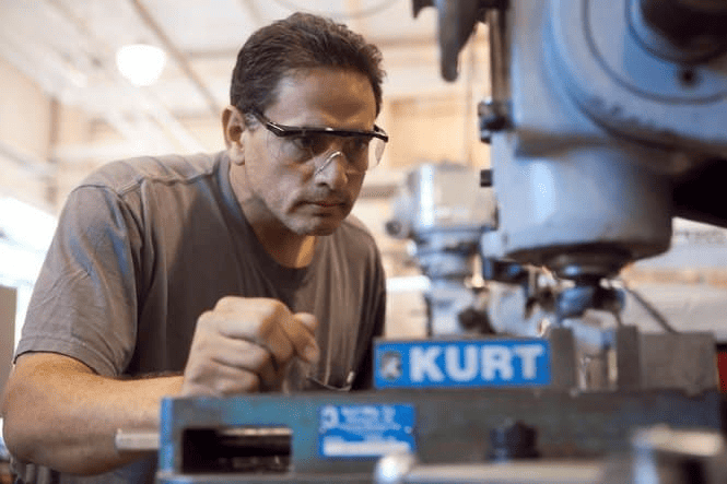 Man with dark hair wearing safety glasses focused on a KURT machine in a well-lit workshop.