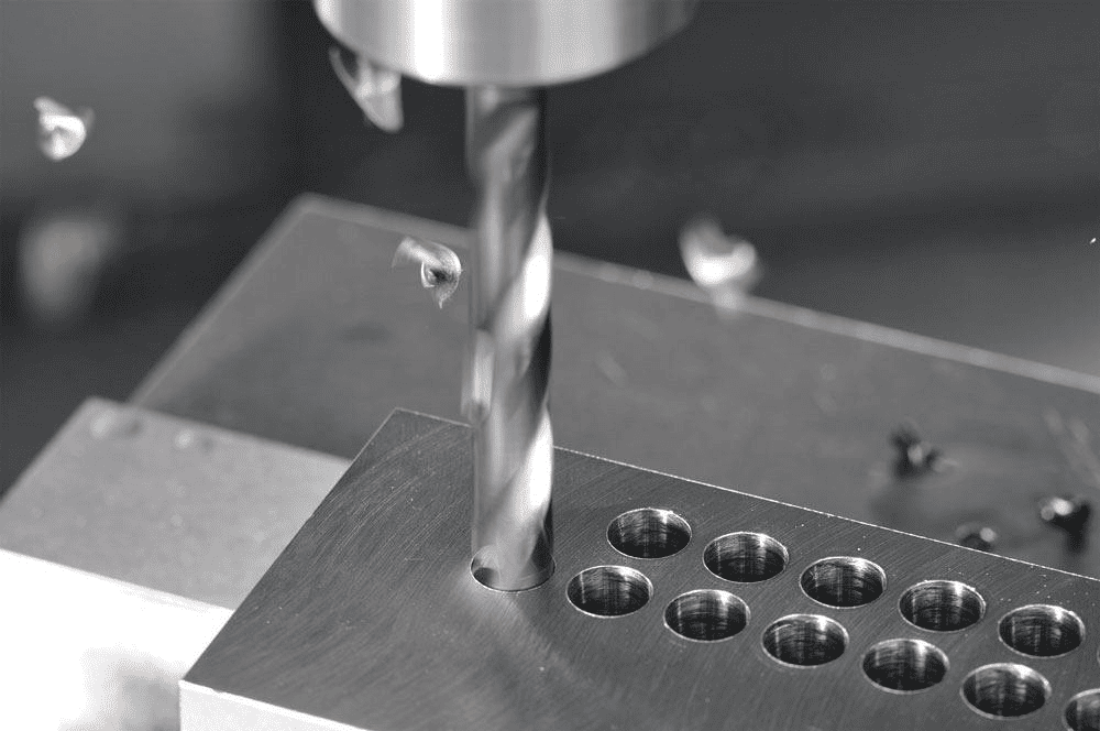 A close-up image of a drill bit or end mill cutting into a metal plate with multiple pre-drilled holes.