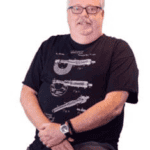 Man with short white hair wearing black shirt with handgun illustrations.