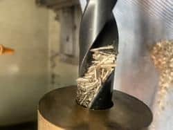 A close-up image of a drill bit machining bronze, with visible cutting edge and accumulated swarf.