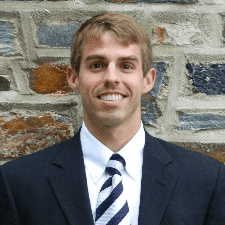 Young man in dark suit standing in front of grayish-brown brick wall.